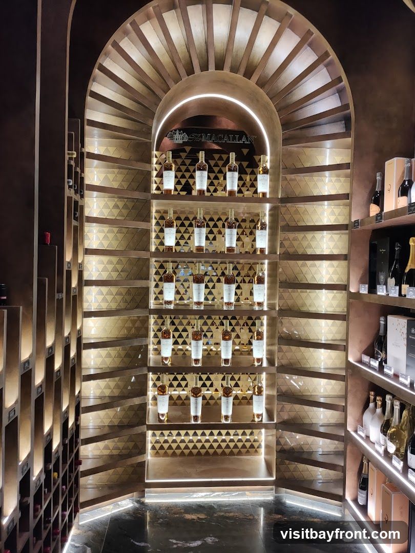 A well-lit display of Macallan whiskey bottles on tiered shelves within a store. The bottles are arranged in rows, with some shelves featuring decorative geometric patterns. The Macallan logo is prominently displayed above the central shelves. Additional bottles and boxes are visible on shelves to the right.