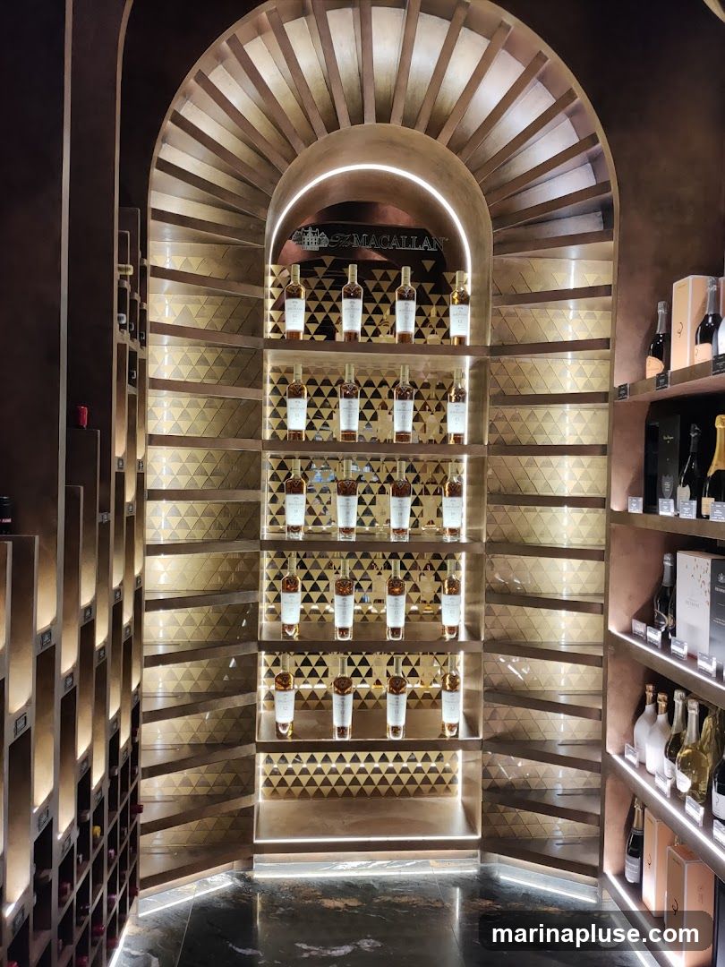 A well-lit display of Macallan whiskey bottles on tiered shelves within a store. The bottles are arranged in rows, with some shelves featuring decorative geometric patterns. The Macallan logo is prominently displayed above the central shelves. Additional bottles and boxes are visible on shelves to the right.