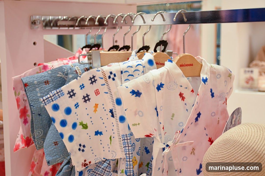 A rack of children's garments, possibly kimonos or yukatas, with various playful patterns including goldfish, flowers, and abstract designs. The garments are displayed on hangers, with some featuring the 'miki house' logo on the wooden hangers. A straw hat is visible on the right side.