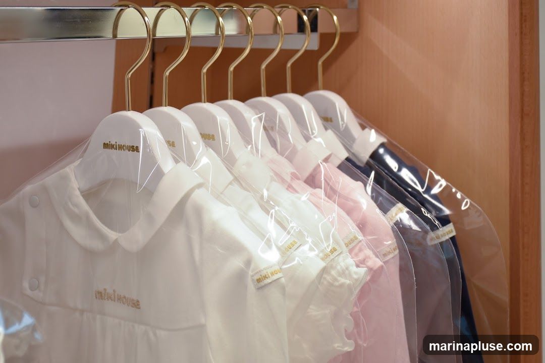 A row of children's clothing items, including onesies and dresses, hang on white hangers with gold hooks. Each garment is individually wrapped in clear plastic. The brand name "miki house" is visible in gold lettering on the hangers and embroidered on some of the clothing. The clothes are in white, pink, and navy blue.
