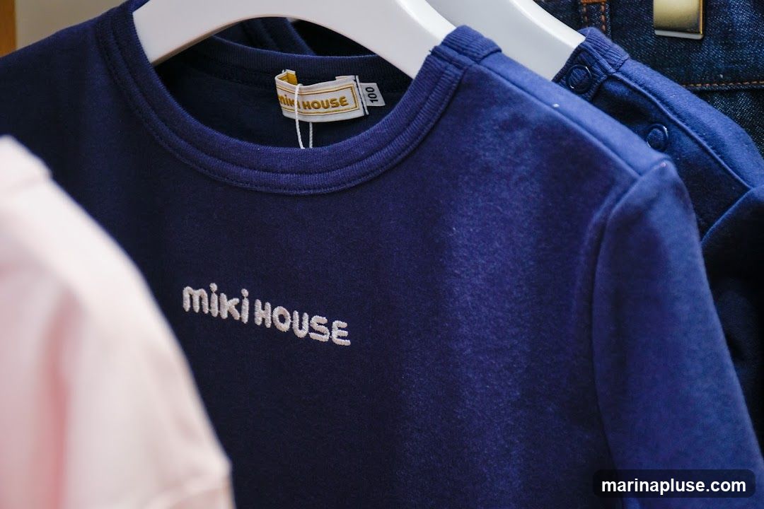 A close-up shot of a navy blue Miki House t-shirt with the brand name embroidered in white. A pink garment is visible on the left, and a denim garment is on the right. A size tag reading '100' is attached to the t-shirt.