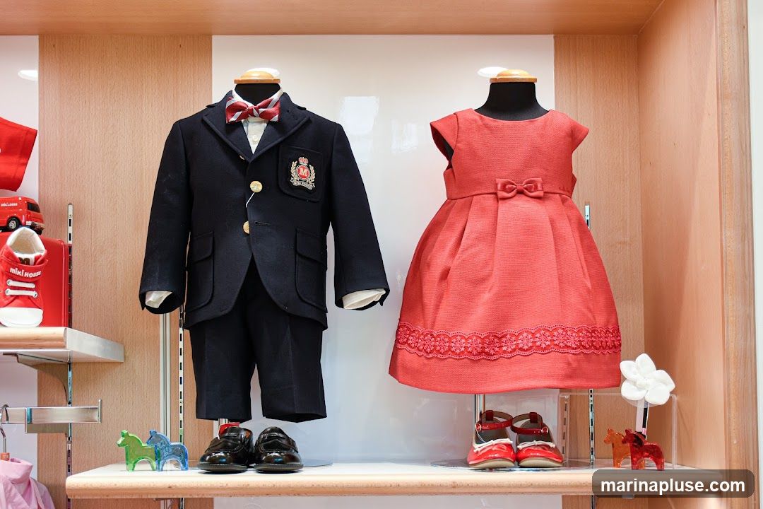A display of children's formal wear includes a navy blue suit with a red and white striped bow tie on a mannequin, and a coral-colored dress with a bow on another mannequin. Both outfits are displayed with matching shoes. To the left, red sneakers with the logo 'miki house' are visible on a shelf, along with a toy red car. Decorative horse figurines in green, blue, orange, and red are also part of the display.