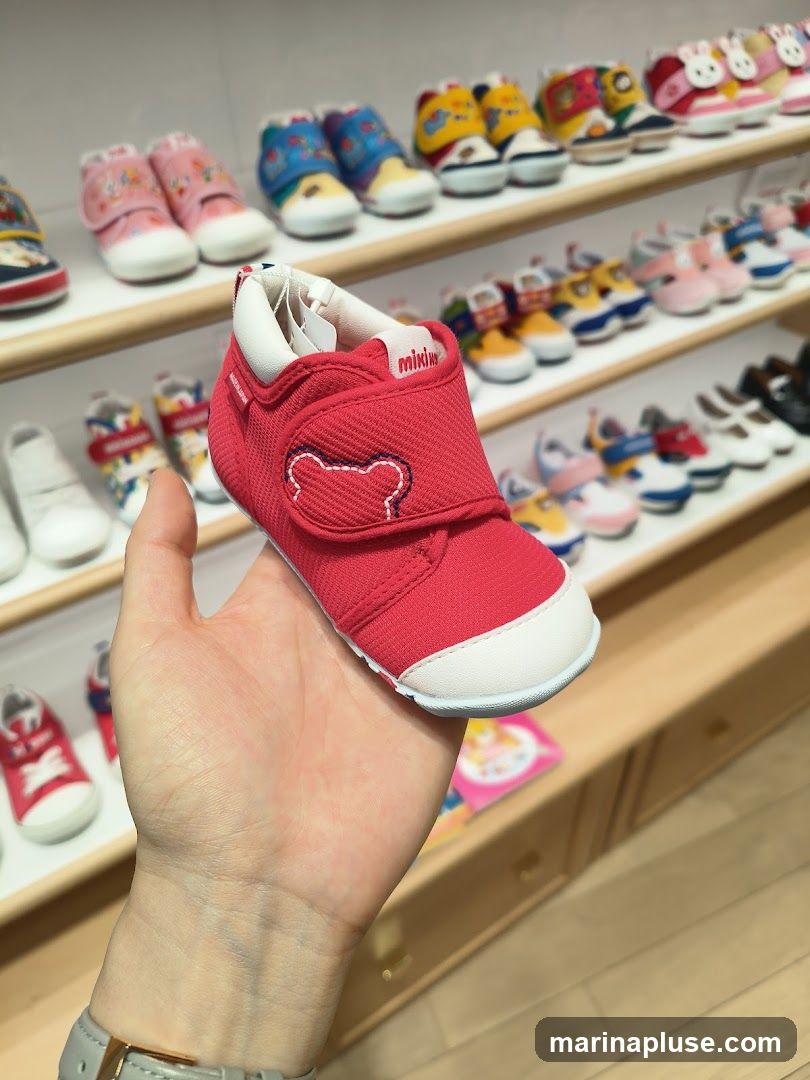 A hand is holding a red baby shoe with white accents and a bear design. In the background, shelves are filled with various colorful baby shoes.