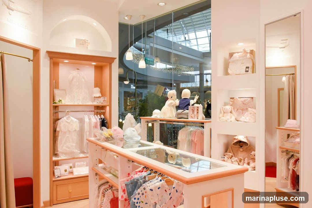 The interior of a brightly lit children's clothing store. Display shelves and racks showcase a variety of baby and children's apparel, including dresses, outfits, and accessories. Mannequins of a boy and girl are positioned near a large window overlooking a mall interior. Stuffed animals and gift boxes are also on display.