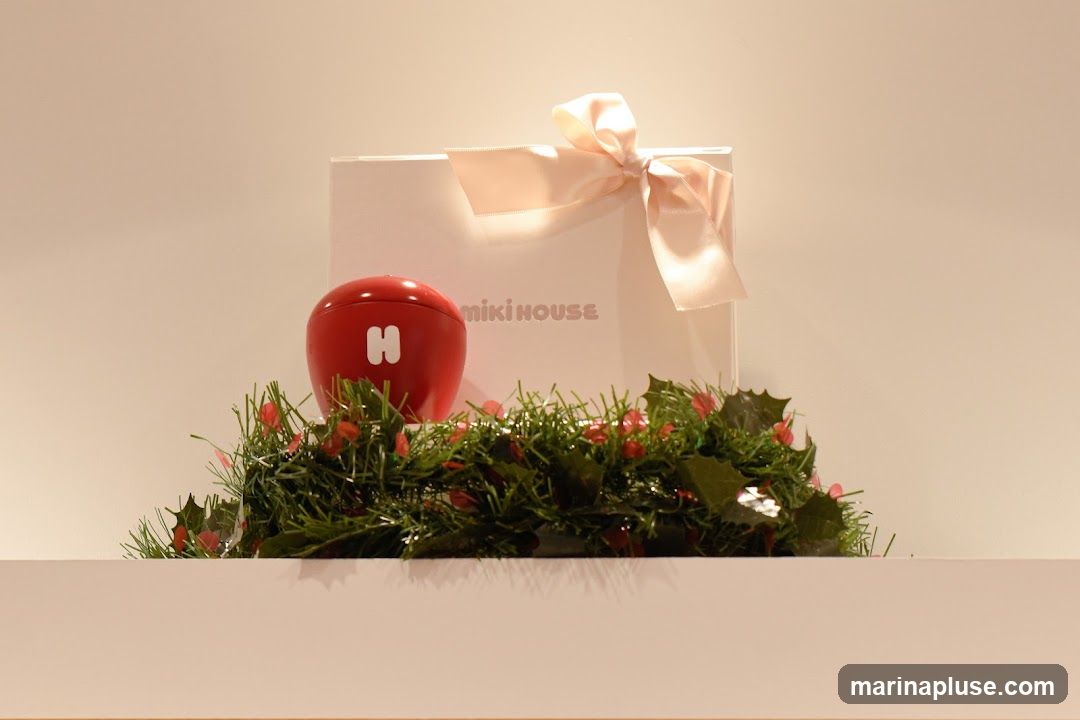 A white gift box with a pink ribbon is displayed behind a red, apple-shaped object with a white 'H' on it. The items are placed on a shelf in front of a plain, light-colored background. A garland of green pine needles and red berries encircles the base of the display, suggesting a festive or holiday theme. The text 'miki HOUSE' is visible on the gift box.