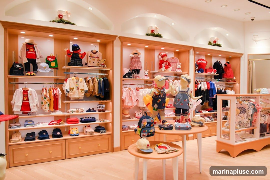 A brightly lit interior of a children's clothing store, showcasing apparel on mannequins and shelves. The store features wooden shelving units displaying a variety of baby and children's clothes, including outfits, jackets, shirts, and pants. Mannequins are dressed in colorful ensembles. A display case on the right exhibits more clothing items. The overall ambiance is clean and organized, with a focus on children's fashion.