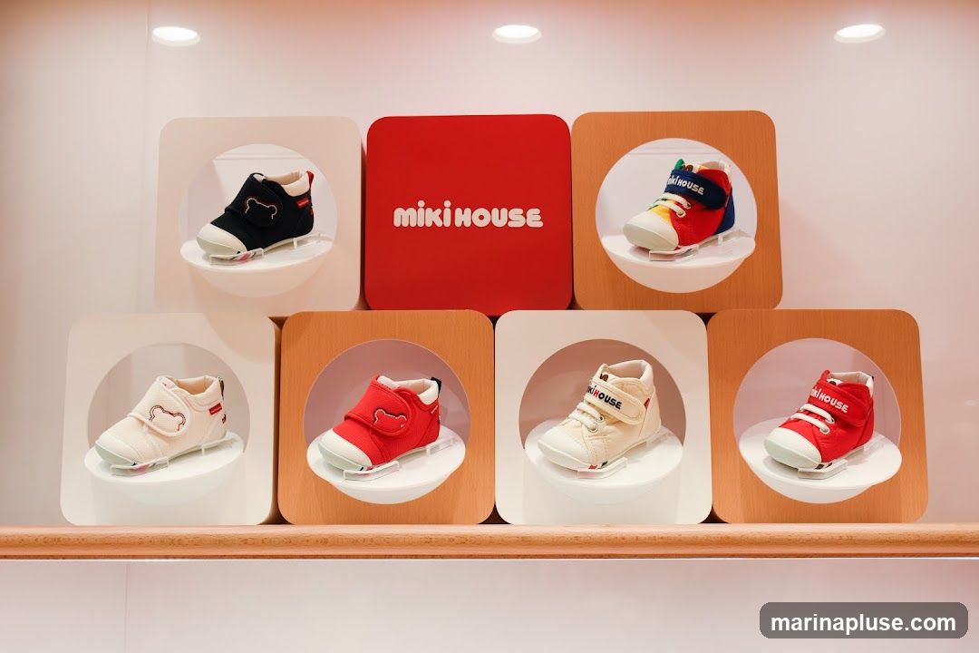 A display of Miki House children's shoes on shelves. The shoes are presented in individual white and wooden framed circular cutouts. A prominent red square with the Miki House logo is in the center of the top row. The shoes are in various colors including black, red, white, and multi-colored.