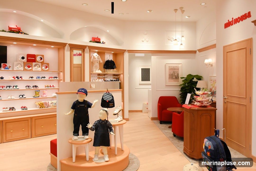 The interior of a brightly lit children's clothing store. On the left, shelves display rows of shoes and boots. In the center, mannequins dressed in denim outfits stand on display platforms. A backpack hangs on a stand behind them. Further back, a seating area with red chairs and a small table is visible, along with a wooden cabinet displaying a tiered stand of packaged goods. A door is visible on the right side of the image. The store's name, "miki house," is displayed in red lettering on the wall above the door.