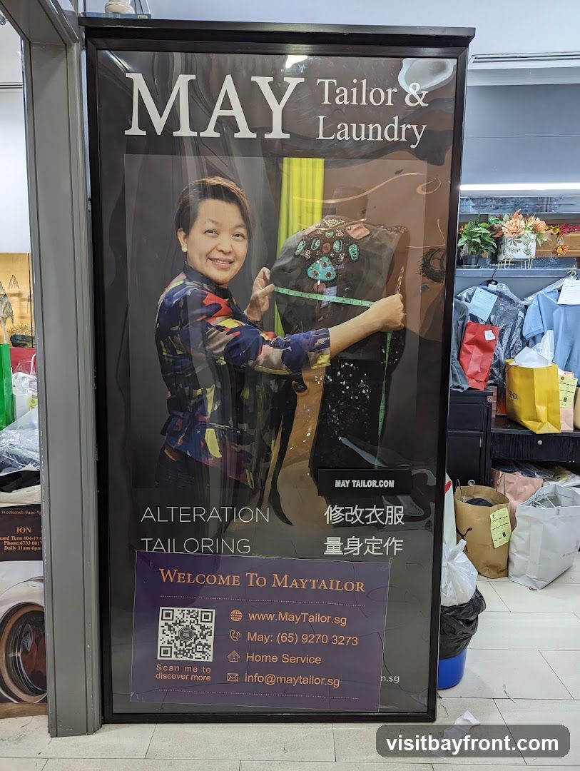 A large advertisement for May Tailor & Laundry features a woman holding a measuring tape against a garment. The text indicates services such as alteration and tailoring. Contact information, including a website, phone number, and email address, is provided. The advertisement is displayed in a frame, likely in a public area or shop.
