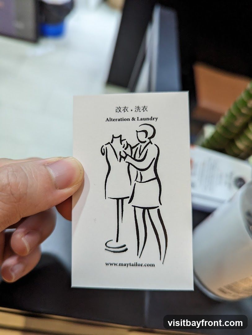 A business card with a line drawing of a tailor fitting a garment on a mannequin. The text reads "改衣・洗衣 Alteration & Laundry" and a website address "www.maytailor.com". The card is held by a person's thumb and index finger. The background is blurred and appears to be an indoor setting with office-like furniture and a plant.