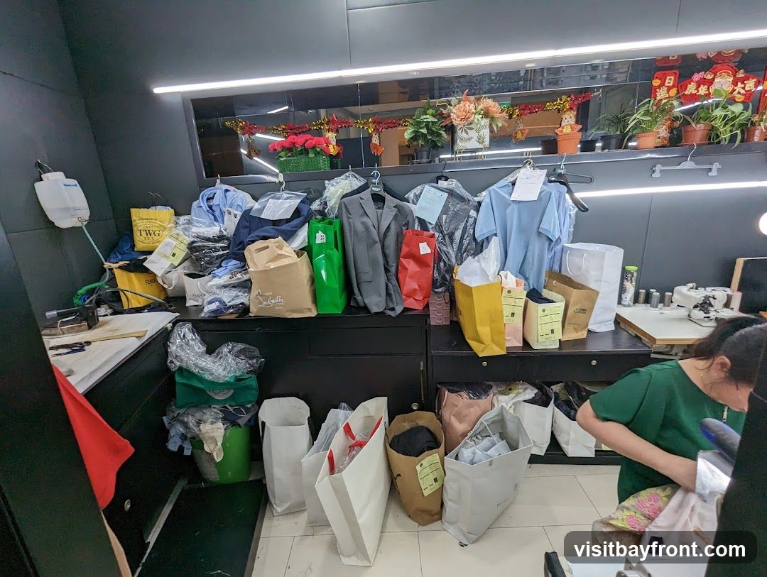 A cluttered backroom of a clothing store with various garments, shopping bags, and supplies. Clothes are hung on racks and piled on surfaces. Several shopping bags from brands like TWG and Fendi are visible. A sewing machine and ironing board suggest alteration or repair services. A woman in a green shirt is seen working in the foreground. Decorative items and plants are displayed on a shelf above.