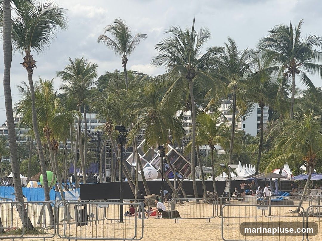 A beach scene with numerous palm trees in the foreground and midground. Behind the trees, a large building complex is visible. In the midground, there is a stage setup with lighting rigs and large screens. Several tents and umbrellas are set up on the sand, with people relaxing on lounge chairs. Metal barriers are placed on the sand, separating areas of the beach. The sky is overcast.