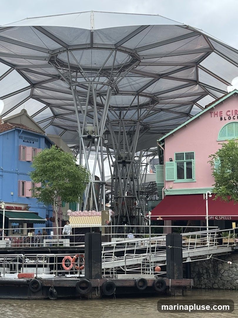 A view of colorful buildings along a riverfront, with a large, modern canopy structure overhead. A dock with a ramp extends into the water. The pink building on the right has a sign that reads "THE CIRCLE" and another sign below a red awning says "KOPI & SPELLS". The blue building on the left has a sign that says "THE RIVERFRONT BLOCK B".