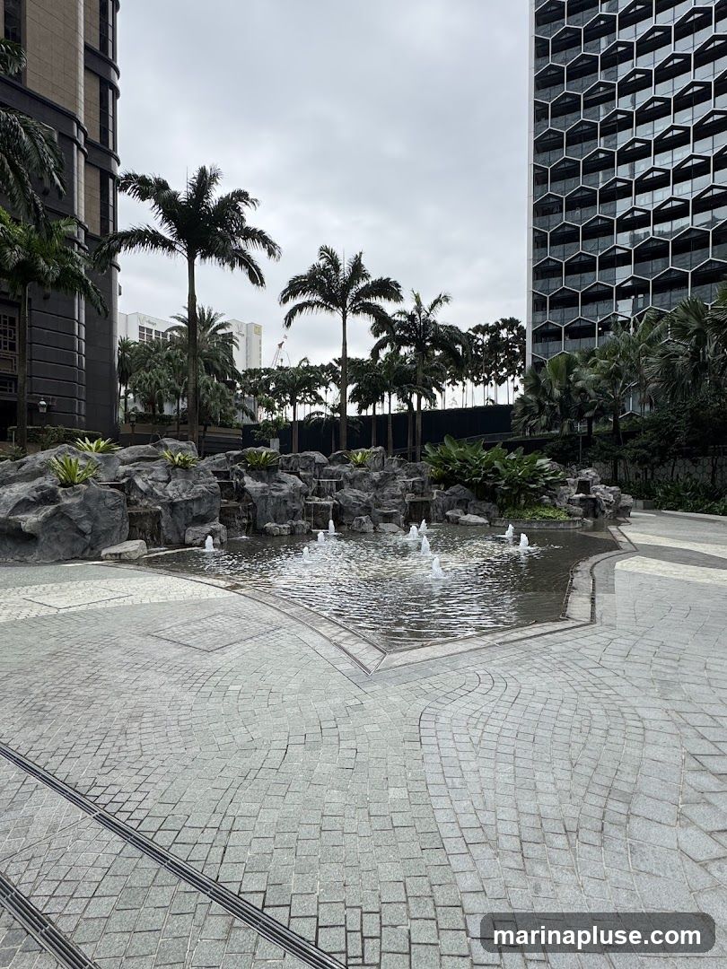 A serene outdoor scene featuring a multi-tiered fountain with several spouts of water cascading over rocks. The fountain is surrounded by lush greenery and palm trees. In the background, two distinct modern buildings stand tall against an overcast sky. The building on the left has a darker, textured facade, while the building on the right features a striking honeycomb-patterned glass exterior. The foreground is paved with intricate stone tiling, creating a clean and contemporary aesthetic.