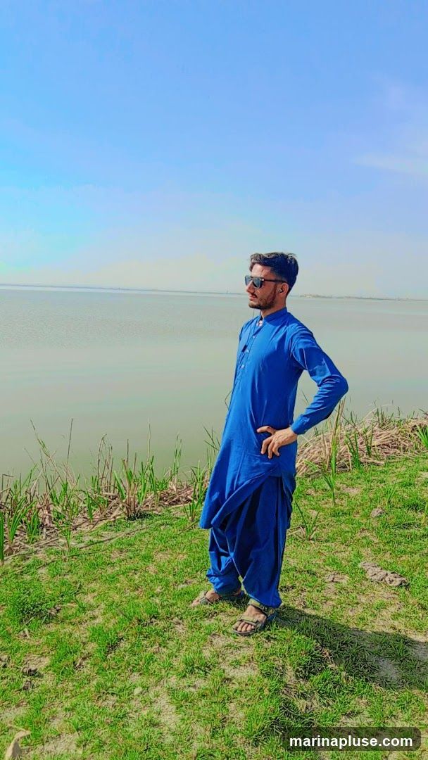 A man wearing a blue shalwar kameez and sunglasses stands on a grassy bank, looking out at a body of water under a clear blue sky.