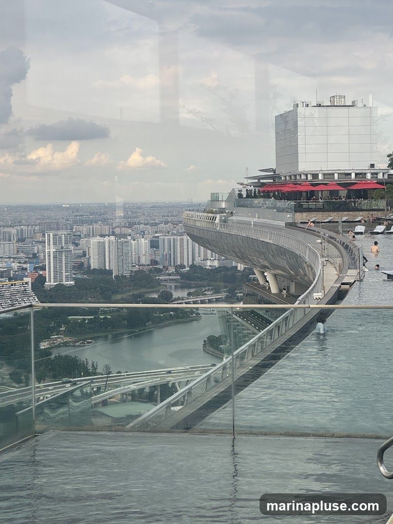 An infinity pool with clear blue water is in the foreground, with a glass railing. Beyond the pool, a curved walkway with red umbrellas and seating areas is visible, with people relaxing. In the background, a dense cityscape stretches to the horizon under a cloudy sky. Reflections of trees and buildings are visible on the glass surfaces.