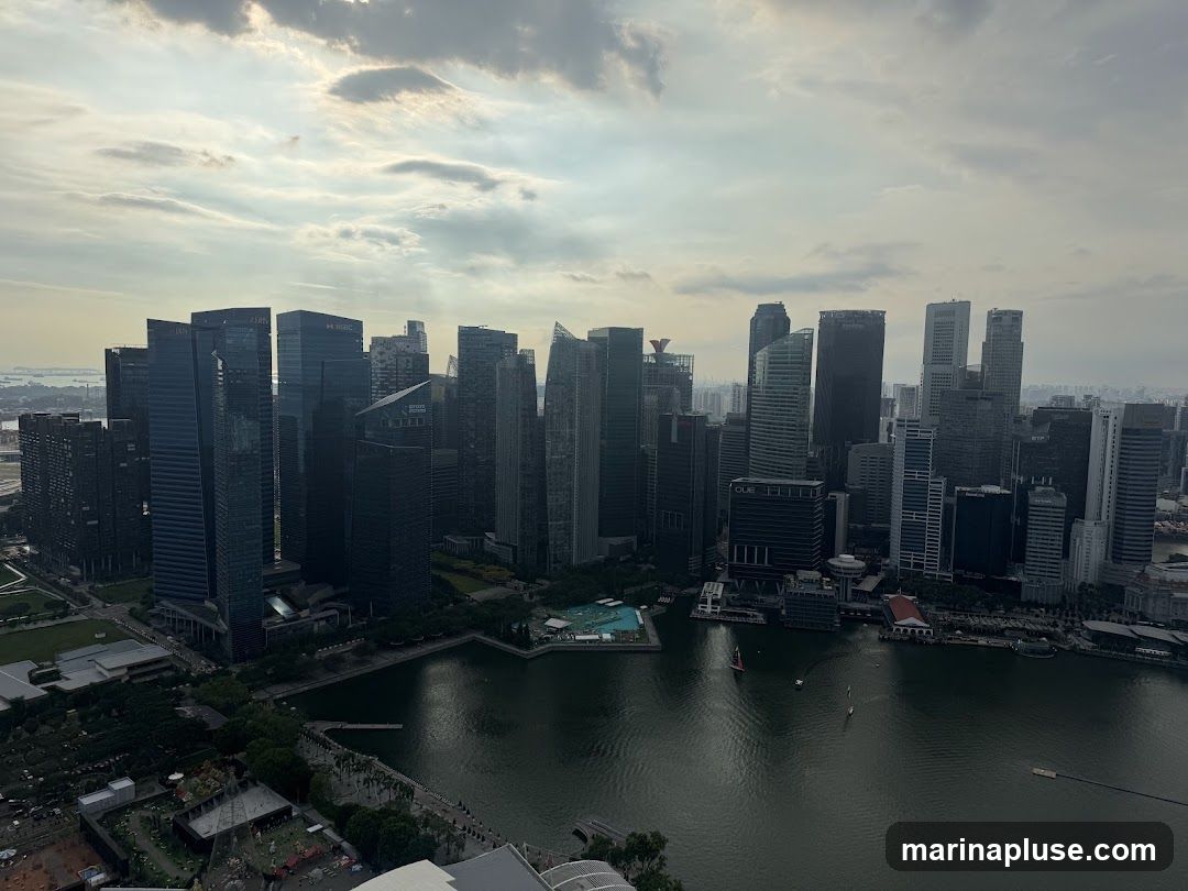 An aerial view of a dense city skyline with numerous skyscrapers of varying heights and architectural styles. The buildings are clustered together, with some appearing to be office buildings or residential towers. In the foreground, a body of water, possibly a bay or river, reflects the overcast sky. Several boats and yachts are visible on the water. The overall scene depicts a bustling urban environment with a mix of modern architecture and natural elements.