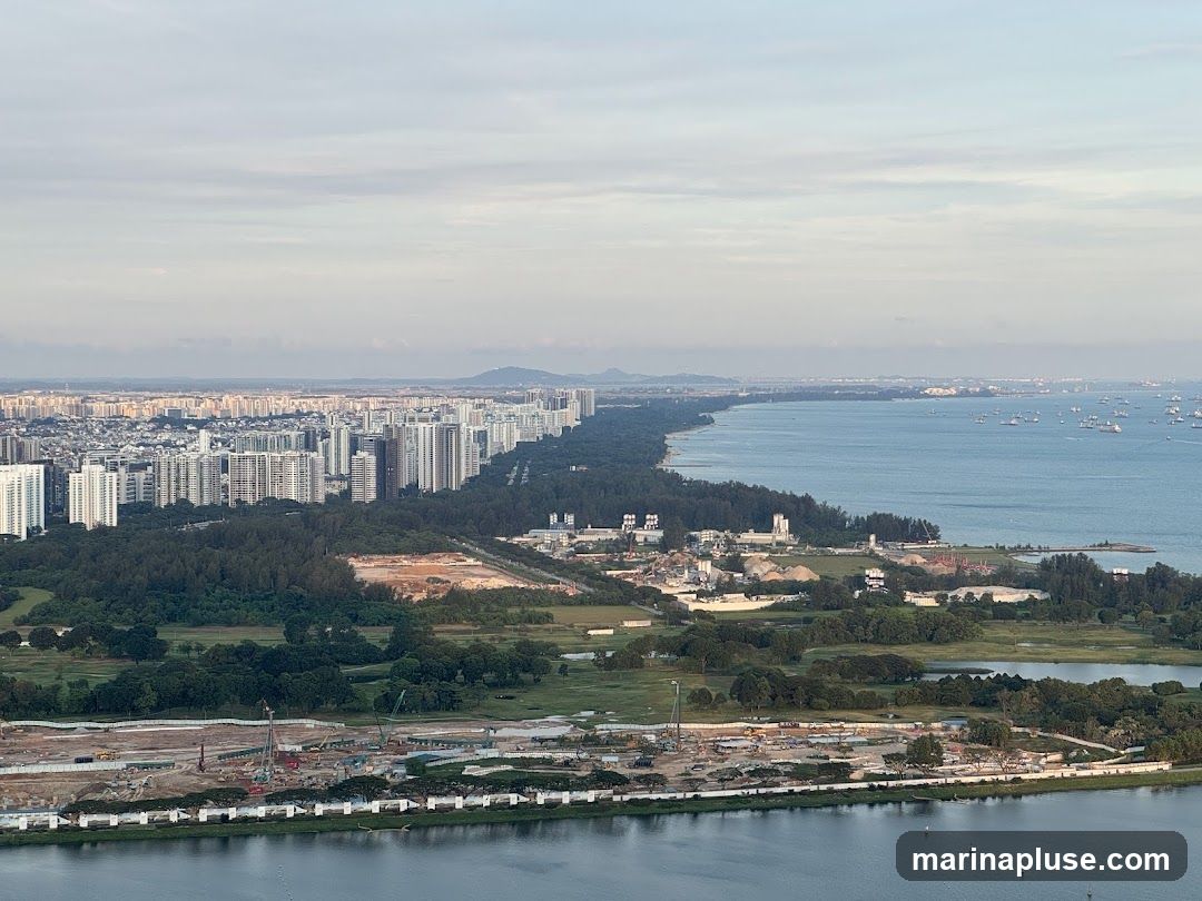An aerial view of a sprawling coastal city with dense residential buildings lining the shore. A large body of water, likely a bay or sea, is visible on the right, dotted with numerous ships. The foreground shows a significant construction site with cranes and earthmoving equipment, indicating ongoing development. Green spaces and trees are interspersed between the urban areas and the construction zone. The sky is partly cloudy with a soft, diffused light.