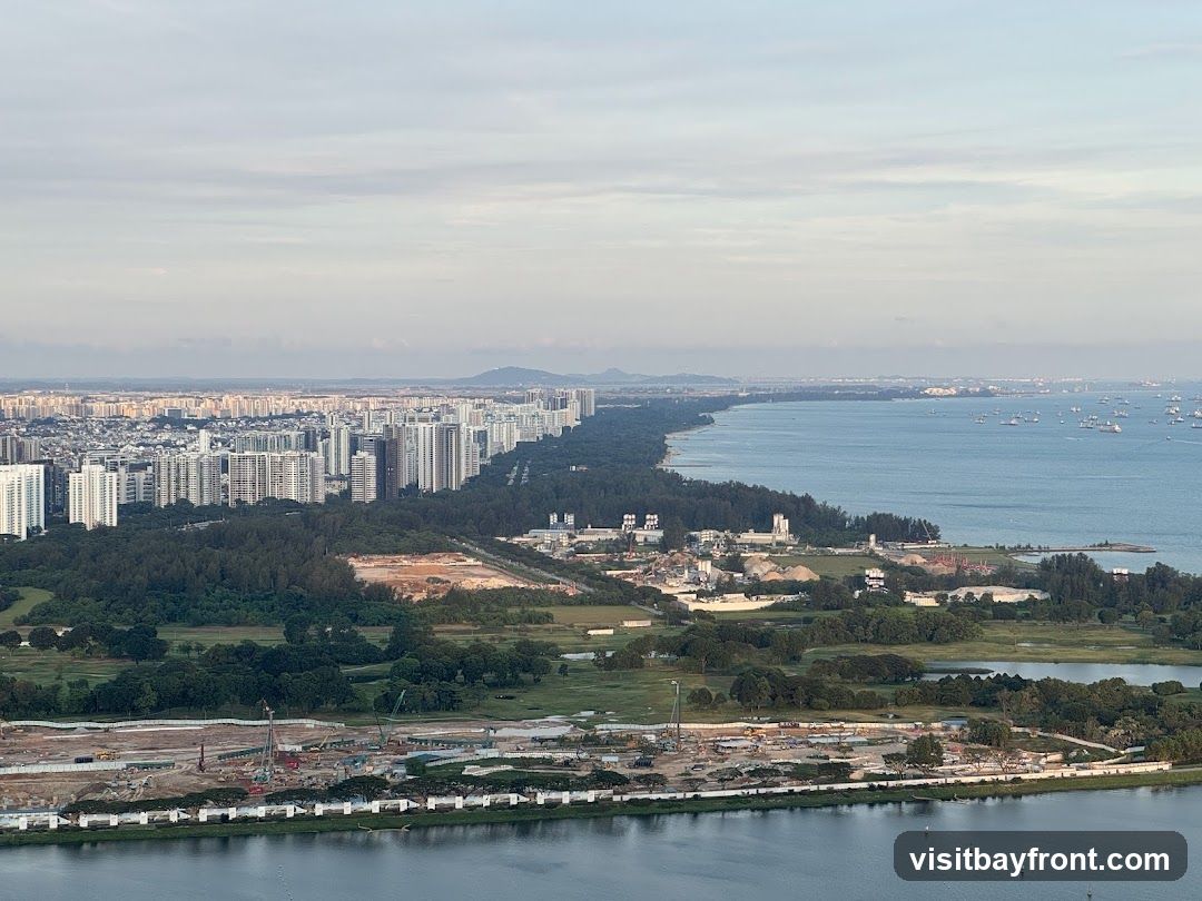 An aerial view of a sprawling coastal city with dense residential buildings lining the shore. A large body of water, likely a bay or sea, is visible on the right, dotted with numerous ships. The foreground shows a significant construction site with cranes and earthmoving equipment, indicating ongoing development. Green spaces and trees are interspersed between the urban areas and the construction zone. The sky is partly cloudy with a soft, diffused light.