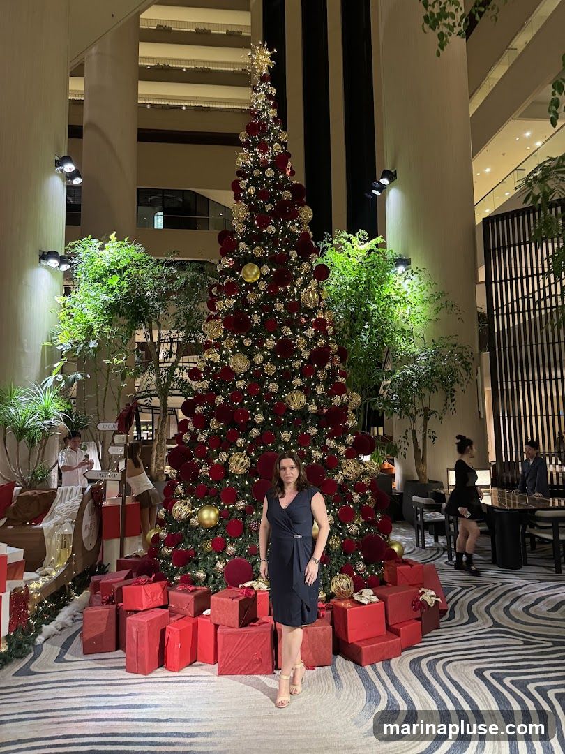 A woman in a dark blue dress stands in front of a large, elaborately decorated Christmas tree. The tree is adorned with red and gold ornaments, lights, and a star topper. It is surrounded by red gift boxes. The scene appears to be set in a hotel lobby or a similar indoor public space, with lush greenery and modern architectural elements visible in the background. Other people are seen in the distance, and the floor has a distinctive wavy pattern.