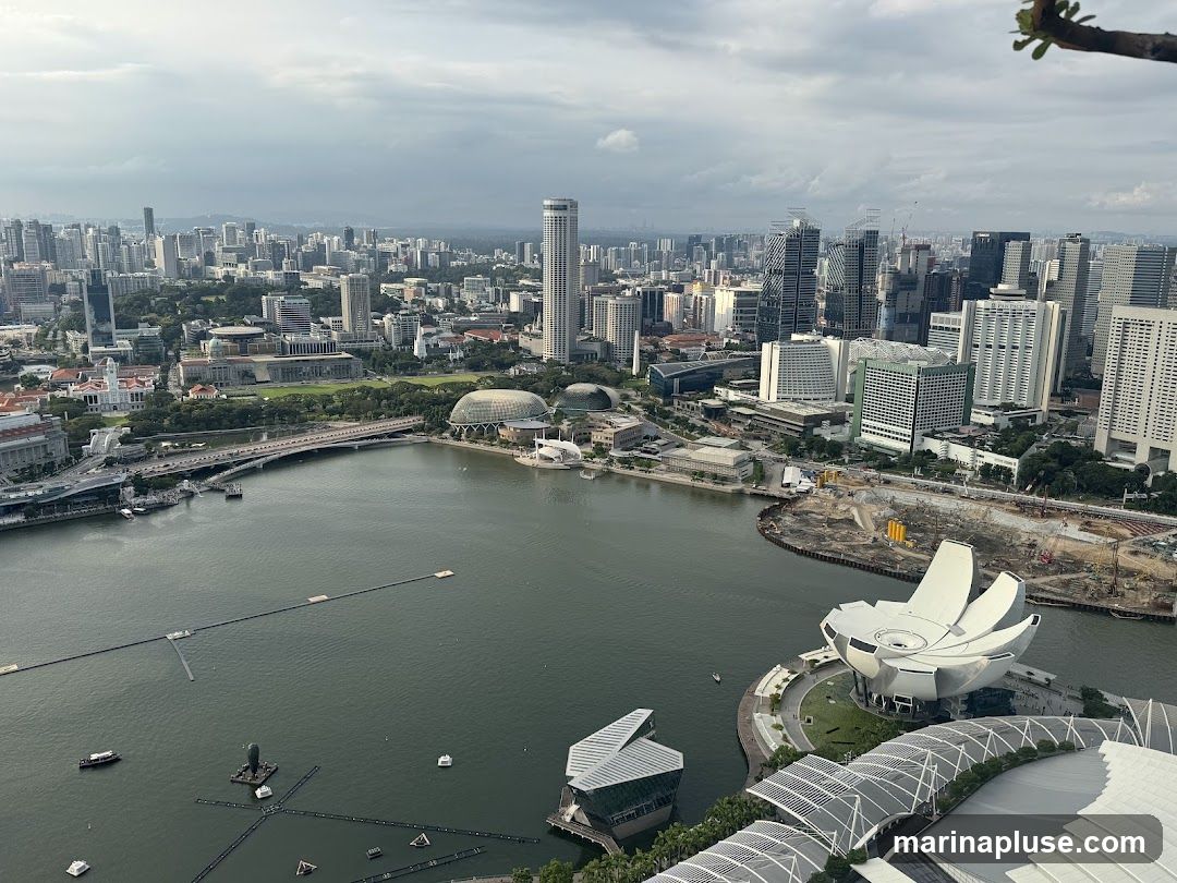 An aerial view of the Singapore skyline, featuring numerous skyscrapers and the Marina Bay area. The image captures the city's dense urban landscape, with a large body of water in the foreground. Notable structures include the ArtScience Museum with its distinctive lotus-like design, the Esplanade – Theatres on the Bay, and various bridges and buildings lining the waterfront. The sky is overcast, suggesting a cloudy day. Construction is visible in some areas, indicating ongoing development.