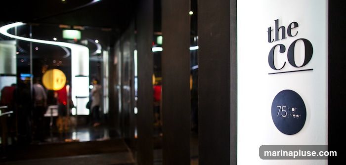A close-up view of a white wall with black lettering that reads "the CO" and a circular emblem below it with "75" and "street" inside. To the left, the blurred interior of a modern establishment with people visible.