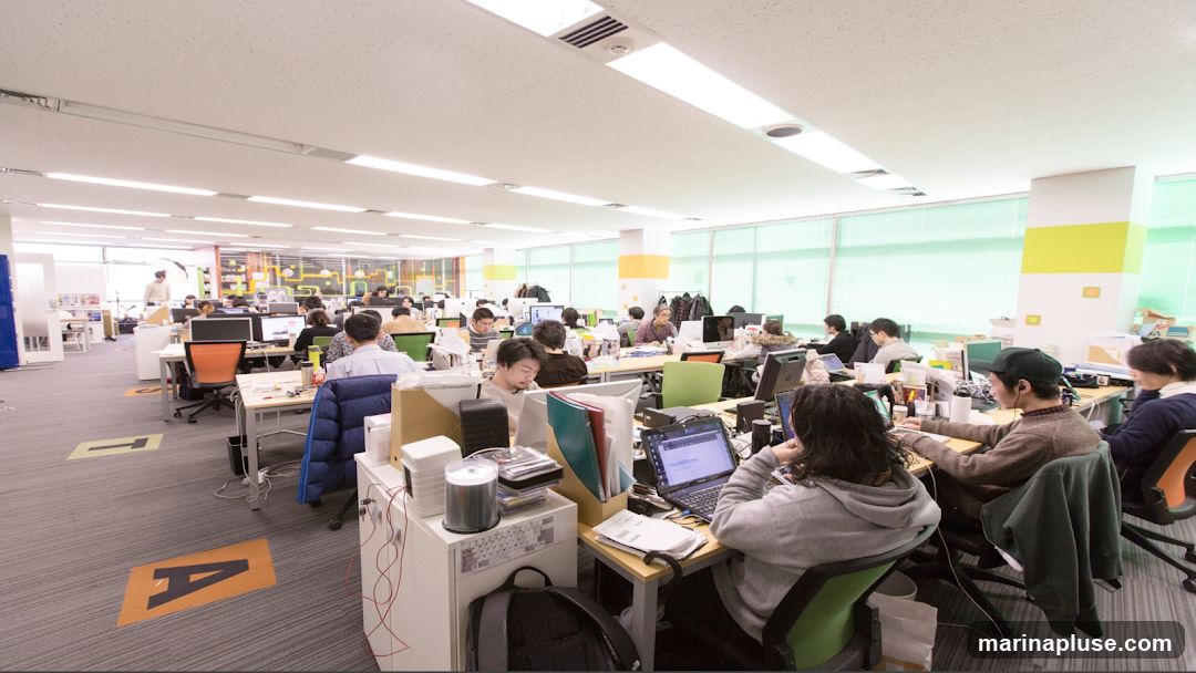 An open-plan office space filled with employees working at their desks. The room is brightly lit with fluorescent lights and has large windows with green blinds. Desks are equipped with computers, monitors, and various office supplies. The carpet has large orange and yellow letter markings.
