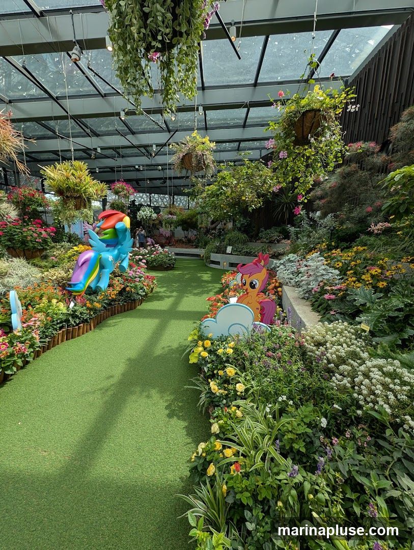 A lush indoor garden with a glass ceiling, featuring a variety of flowering plants and hanging greenery. A pathway made of artificial turf winds through the display. Two colorful My Little Pony figures, Rainbow Dash and Applejack, are placed among the flowers, adding a whimsical touch. Several people can be seen in the background, enjoying the garden.