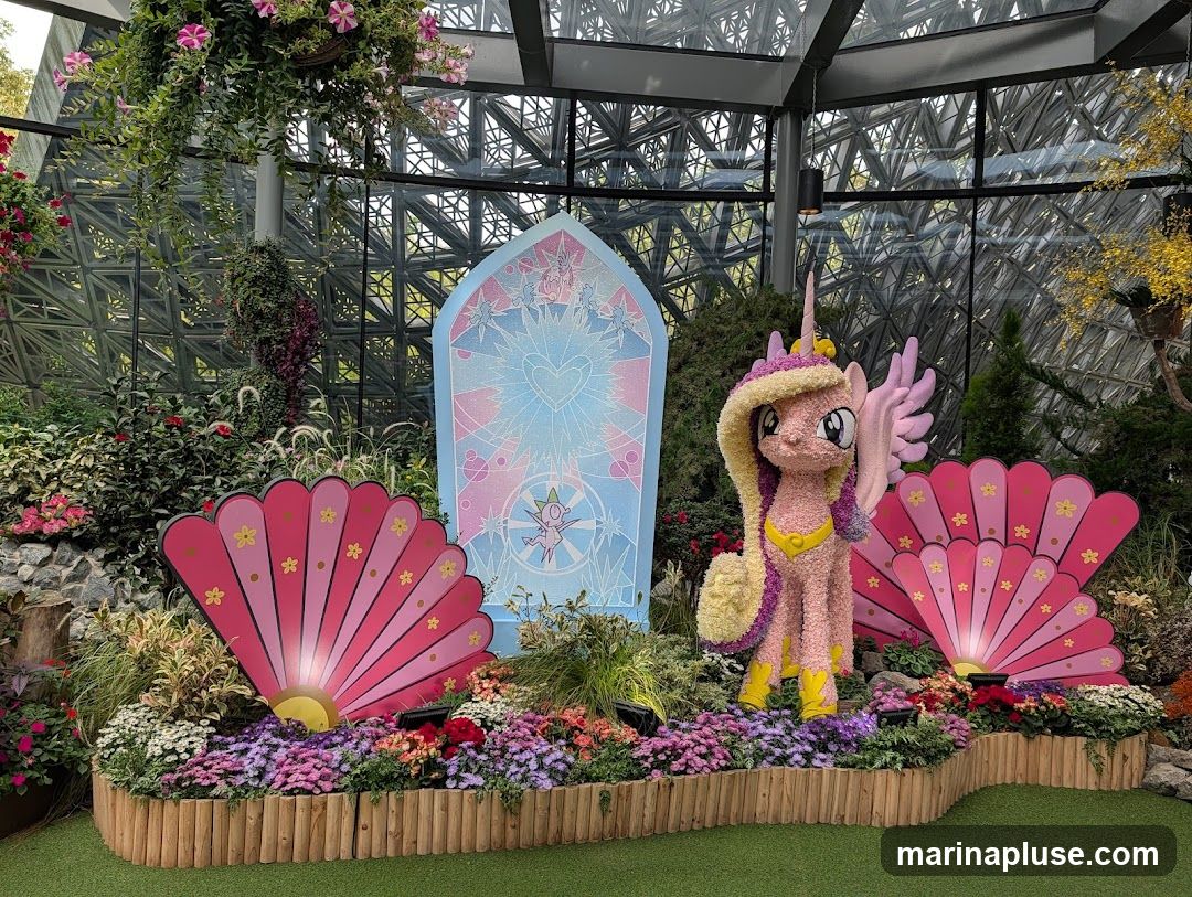 A whimsical floral display featuring a My Little Pony character, possibly Princess Cadance, made of pink and yellow flowers. The character stands between two large pink fan-shaped decorations adorned with smaller yellow flowers. Behind the display is a stained-glass-like panel with a heart and celestial motifs. The entire arrangement is set within a lush garden environment, possibly a conservatory, with various plants, flowers, and a geodesic dome-like ceiling structure visible in the background.
