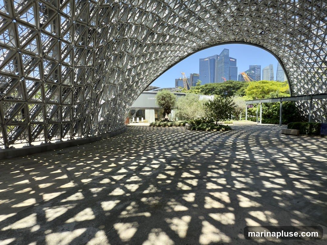 A wide-angle shot captures the intricate geometric metal framework of a modern pavilion, casting dappled shadows on the concrete ground. Beyond the structure, a lush green landscape with trees leads to a backdrop of contemporary skyscrapers and construction cranes under a clear blue sky. The building entrance with the word "GARDEN" is visible in the mid-ground.