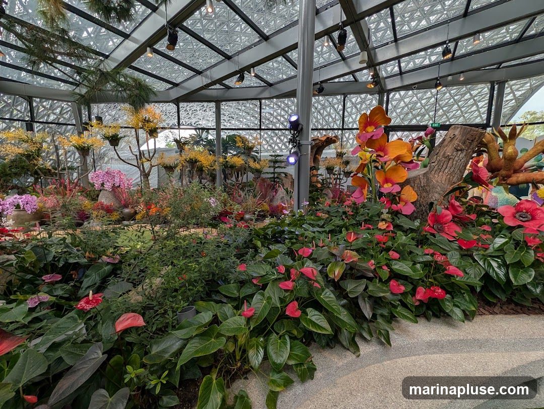 A vibrant indoor botanical display featuring a variety of flowers and plants under a geodesic glass roof. The foreground is filled with lush green foliage and numerous red and pink anthurium flowers. Behind them, a collection of yellow and pink orchids are arranged on branches and in pots. A large, decorative orange and pink orchid bloom is prominently displayed near a rustic wooden log. The background shows more floral arrangements and the intricate structure of the greenhouse ceiling with spotlights.