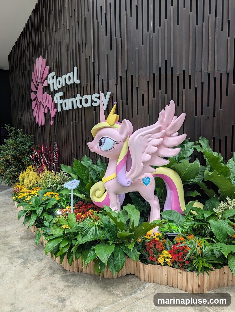 A pink unicorn statue with wings stands in a garden bed filled with colorful flowers and lush green plants. Behind the statue, a wooden wall features the words "Floral Fantasy" in large, white lettering, accompanied by a pink floral design.