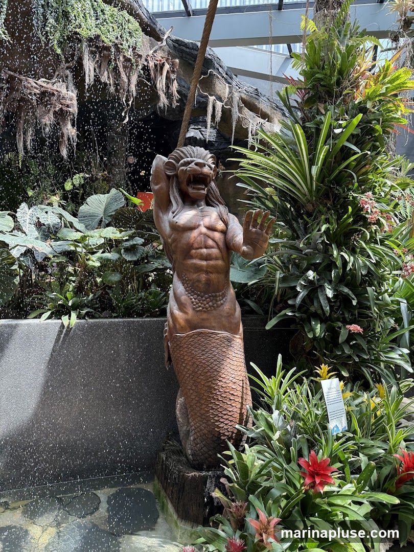 A statue of a mythical creature, resembling a merman with a lion-like head, stands amidst a vibrant and lush garden. The statue is made of a bronze-like material and depicts the creature with its mouth open in a roar and one hand raised. Behind the statue, a waterfall cascades down a rocky surface covered in moss and hanging plants. The surrounding area is filled with a variety of tropical plants, including large-leafed foliage, bromeliads, and flowering plants in shades of red and pink. A small informational sign is visible among the plants on the right side of the image. The scene appears to be indoors, possibly in a botanical garden or conservatory, with a glass ceiling visible in the background.