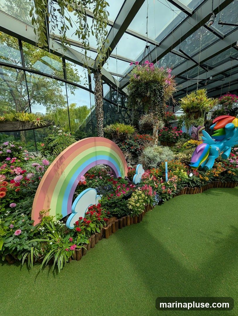A vibrant indoor garden display featuring a variety of colorful flowers, lush greenery, and decorative elements. A large rainbow arch and clouds are prominently placed amidst the plants, with a figurine of a rainbow-maned horse on the right. The space is enclosed by a glass roof and walls, allowing natural light to illuminate the scene.