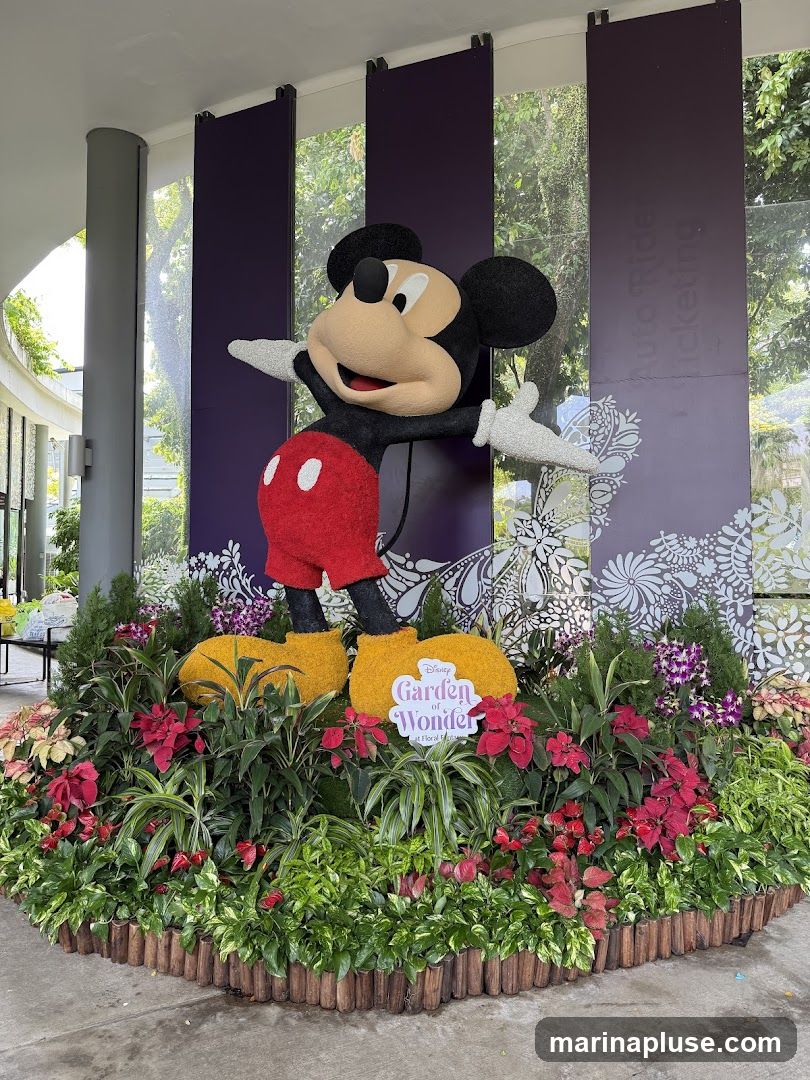 A large, plush Mickey Mouse figure stands in the center of a vibrant floral display. He is surrounded by lush green plants, red poinsettias, and purple orchids. A sign in front of the display reads "Disney Garden of Wonder at Flor de Fes". In the background, tall purple banners with floral patterns are visible, along with trees and foliage through a glass wall.