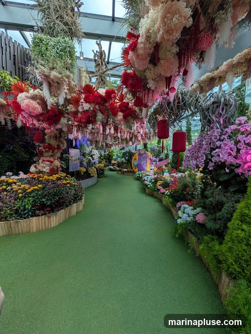 A winding green pathway leads through a vibrant floral display. Overhead, a canopy of dried flowers in shades of red, pink, and white hangs, adorned with red lanterns and delicate tassels. The pathway is bordered by lush flower beds featuring a variety of colorful blooms, including yellow, pink, purple, and blue flowers, as well as green foliage. In the distance, a whimsical purple structure with a yellow roof is visible. The overall scene is a richly decorated indoor garden or conservatory.
