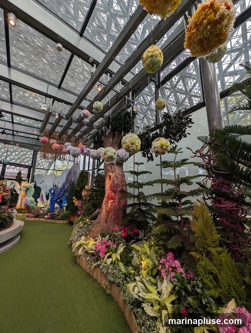 A lush indoor garden display with a geodesic dome ceiling. Hanging floral orbs in various colors adorn the space, suspended from the ceiling. A large, textured tree trunk stands prominently, surrounded by a variety of plants, including flowering bushes and foliage. In the background, colorful statues resembling mythical creatures are visible amidst more greenery. The pathway is covered in green turf.