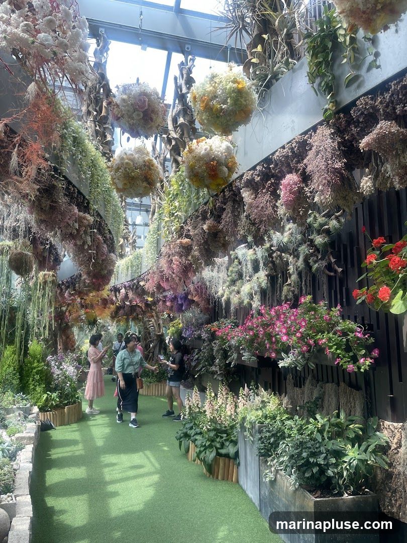 An indoor garden space filled with lush greenery and an abundance of hanging floral arrangements. Various dried and fresh flowers in shades of pink, white, yellow, and orange are suspended from the ceiling, creating a vibrant canopy. The walls are adorned with vertical gardens featuring a variety of plants and flowers. Several people are seen walking along a green pathway, admiring the displays.