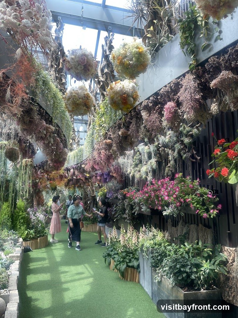 An indoor garden space filled with lush greenery and an abundance of hanging floral arrangements. Various dried and fresh flowers in shades of pink, white, yellow, and orange are suspended from the ceiling, creating a vibrant canopy. The walls are adorned with vertical gardens featuring a variety of plants and flowers. Several people are seen walking along a green pathway, admiring the displays.