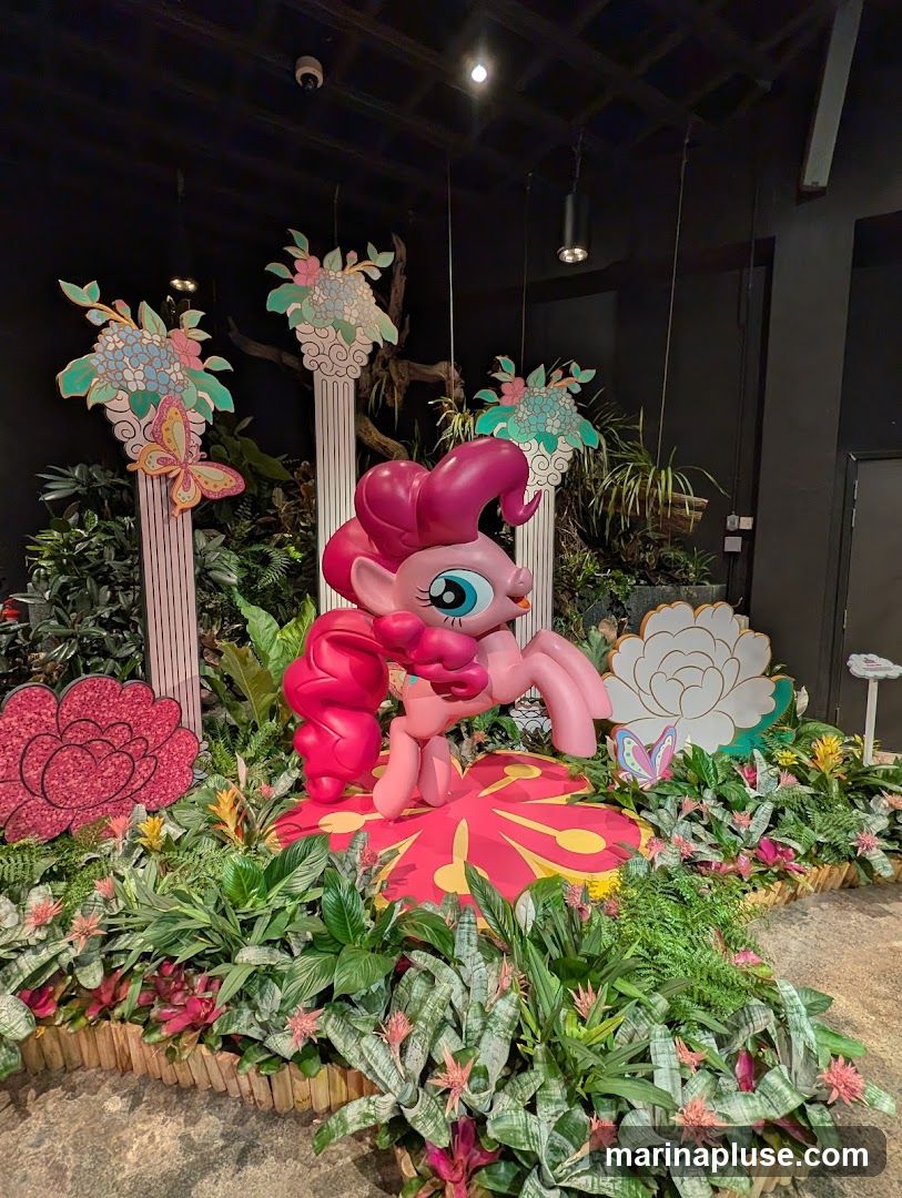 A display featuring a large Pinkie Pie figurine from My Little Pony, surrounded by lush greenery, flowers, and decorative elements like butterflies and floral motifs on pillars.