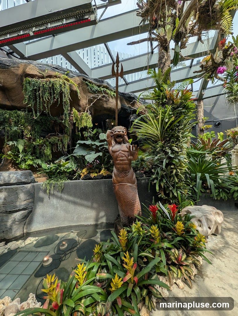 A lush indoor garden features a variety of tropical plants, including bromeliads and ferns, arranged around a small pond. A wooden sculpture of a merman holding a trident stands prominently in the center. The space is covered by a glass ceiling, allowing natural light to filter through.