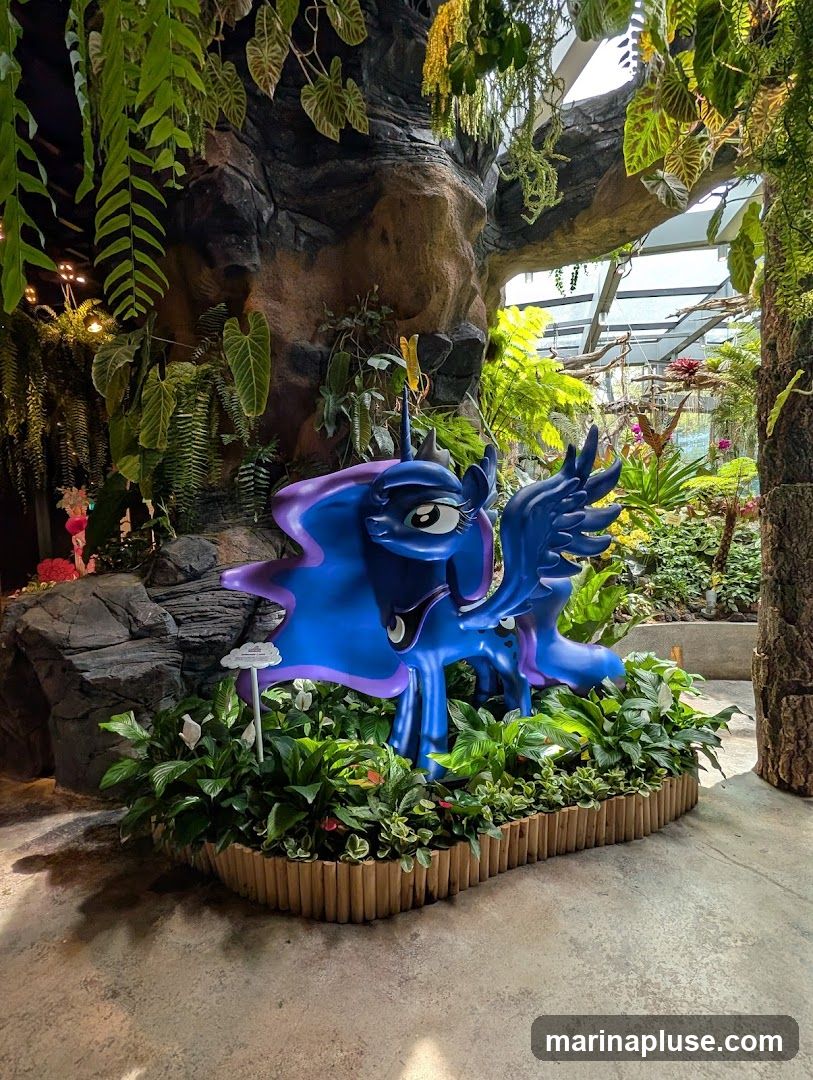 A vibrant blue pony statue, resembling Princess Luna from My Little Pony, is displayed amidst a dense arrangement of green plants. The statue is positioned in front of a large, artificial rock formation covered in hanging ferns and other foliage. The background reveals a greenhouse-like structure with abundant greenery and sunlight filtering through. A small white sign with text is visible near the statue.