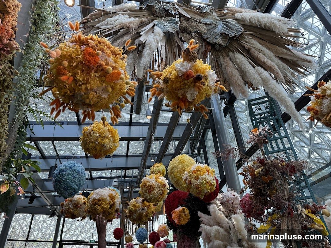A collection of large, spherical floral arrangements made of dried flowers and grasses hang from the ceiling of a conservatory or greenhouse. The arrangements are in various colors, including yellow, orange, blue, and red, and are adorned with smaller dried elements. In the background, a metal ladder and more hanging greenery are visible, along with the geometric pattern of the glass ceiling.