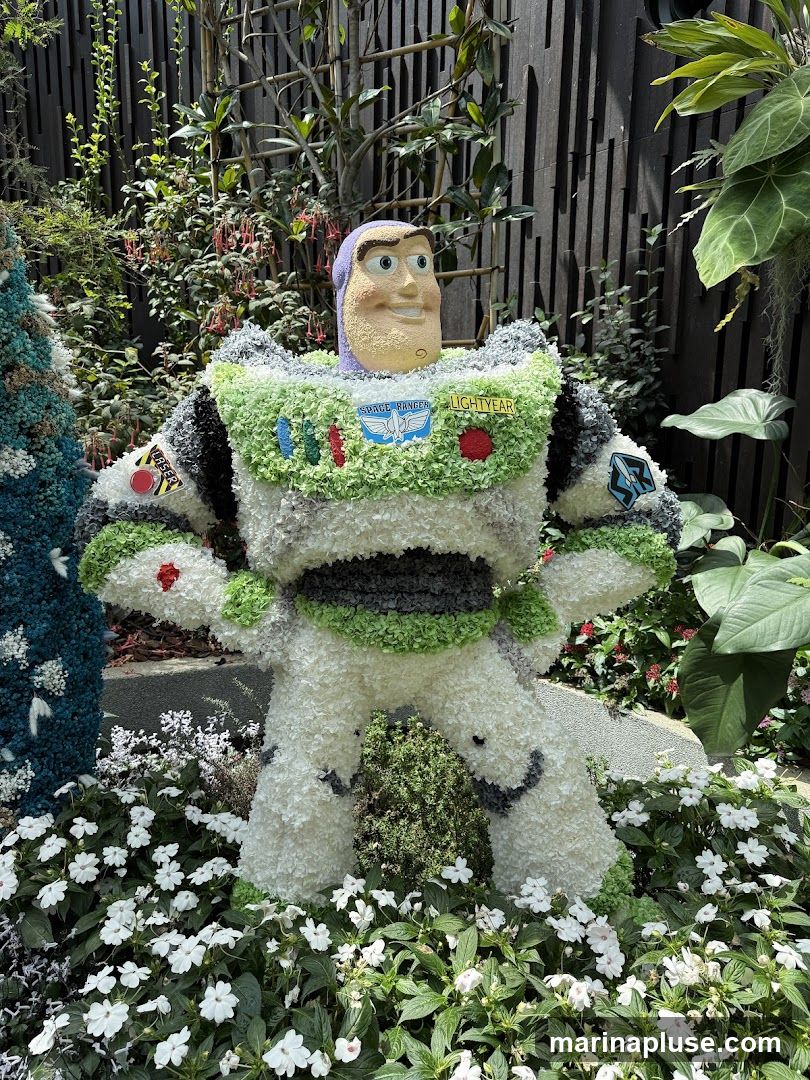 A topiary sculpture of Buzz Lightyear from Toy Story, made entirely of white and green flowers, stands in a garden setting surrounded by lush greenery and white flowers. The sculpture features Buzz's iconic spacesuit with "Space Ranger" and "Lightyear" patches.
