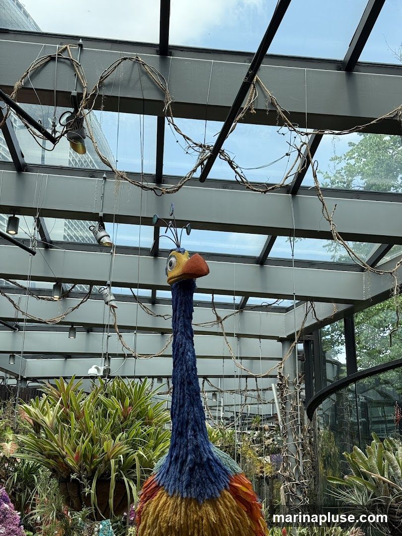 A large, colorful knit sculpture of a bird, resembling Kevin from the movie Up, stands prominently in a greenhouse. The bird has a long blue neck, a yellow and orange body, and a distinctive beak and eyes. It is surrounded by lush green plants, including bromeliads hanging in baskets. The greenhouse has a glass ceiling with metal beams and decorative vines.