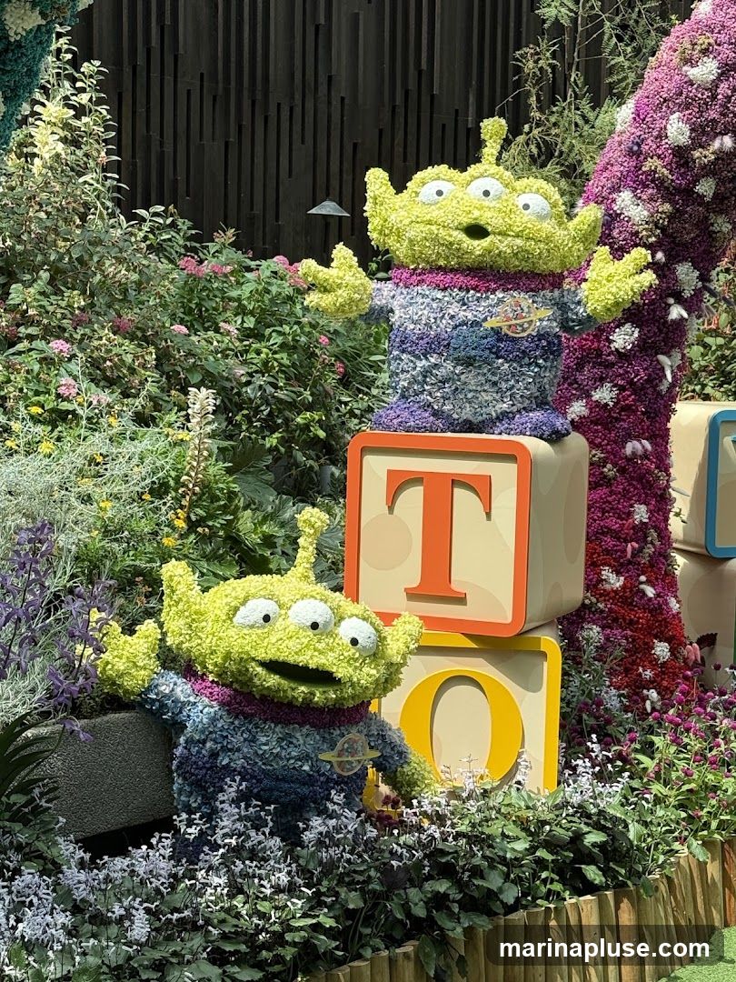 Two floral sculptures of the Toy Story aliens are displayed amongst lush greenery and flowers. The aliens are made of green floral material and wear colorful, striped sweaters. They are positioned in front of large, light-colored blocks with the letters 'T' and 'O' visible. A large, floral archway, predominantly pink and purple, is visible on the right side of the image. The background features a dark, slatted wooden wall.