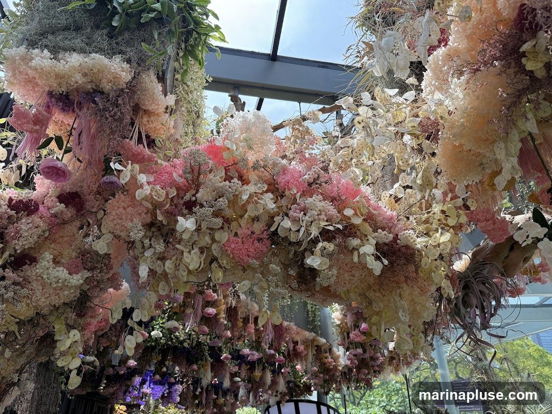 A dense canopy of dried and preserved flowers in shades of pink, cream, and purple hangs from a ceiling structure. The arrangement includes various textures, from delicate, fluffy blooms to larger, rose-like flowers and seed pods. Greenery is also incorporated, with some hanging plants visible. The overall effect is an abundant and whimsical floral display, likely an artistic installation or decoration.