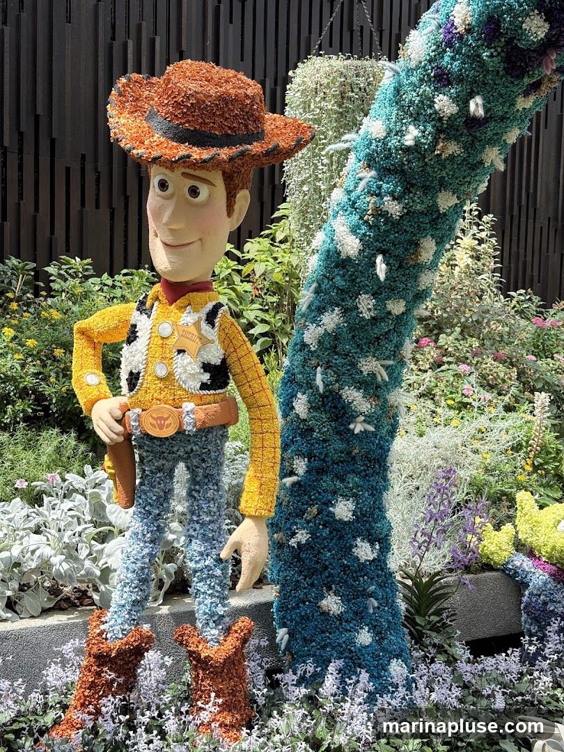 A floral sculpture of Woody from Toy Story stands in a garden setting. The sculpture is made of various textured materials, resembling flowers and foliage. Woody is depicted wearing his signature cowboy hat, vest, shirt, and jeans. A blue and white floral-covered column is to his right. The background features lush greenery and a dark wooden fence.