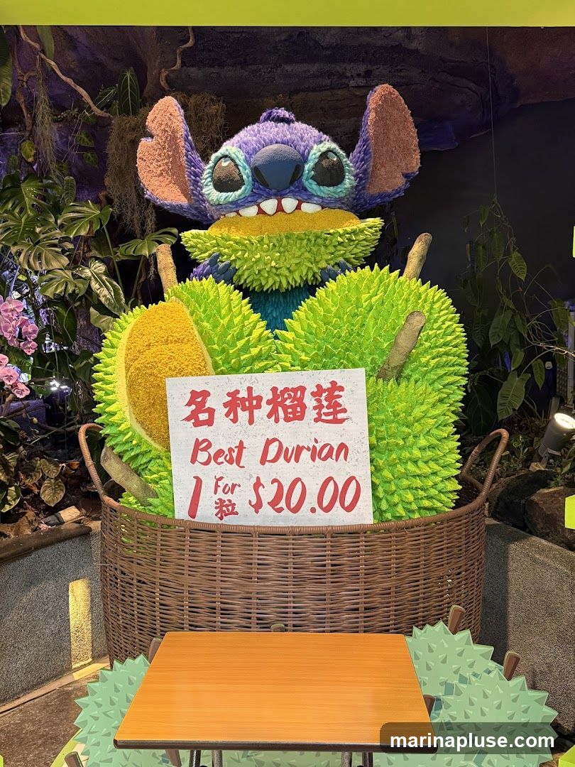 A large, colorful display featuring a character resembling Stitch from Lilo & Stitch, holding a durian. The character is surrounded by large, spiky durian props. A sign in front reads 'Best Durian 1 for $20.00'. A small wooden table is placed in front of the display. The background includes lush greenery and decorative elements, suggesting an indoor market or shop setting.