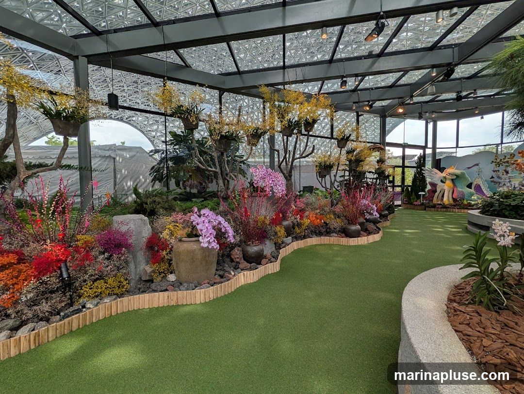 A vibrant indoor garden display featuring a variety of colorful flowers, including orchids, arranged in pots and along a winding path. The space has a glass ceiling and walls, allowing natural light to filter in. In the background, a large sculpture of a winged unicorn and a castle can be seen, suggesting a themed exhibition. The ground is covered with green artificial turf.